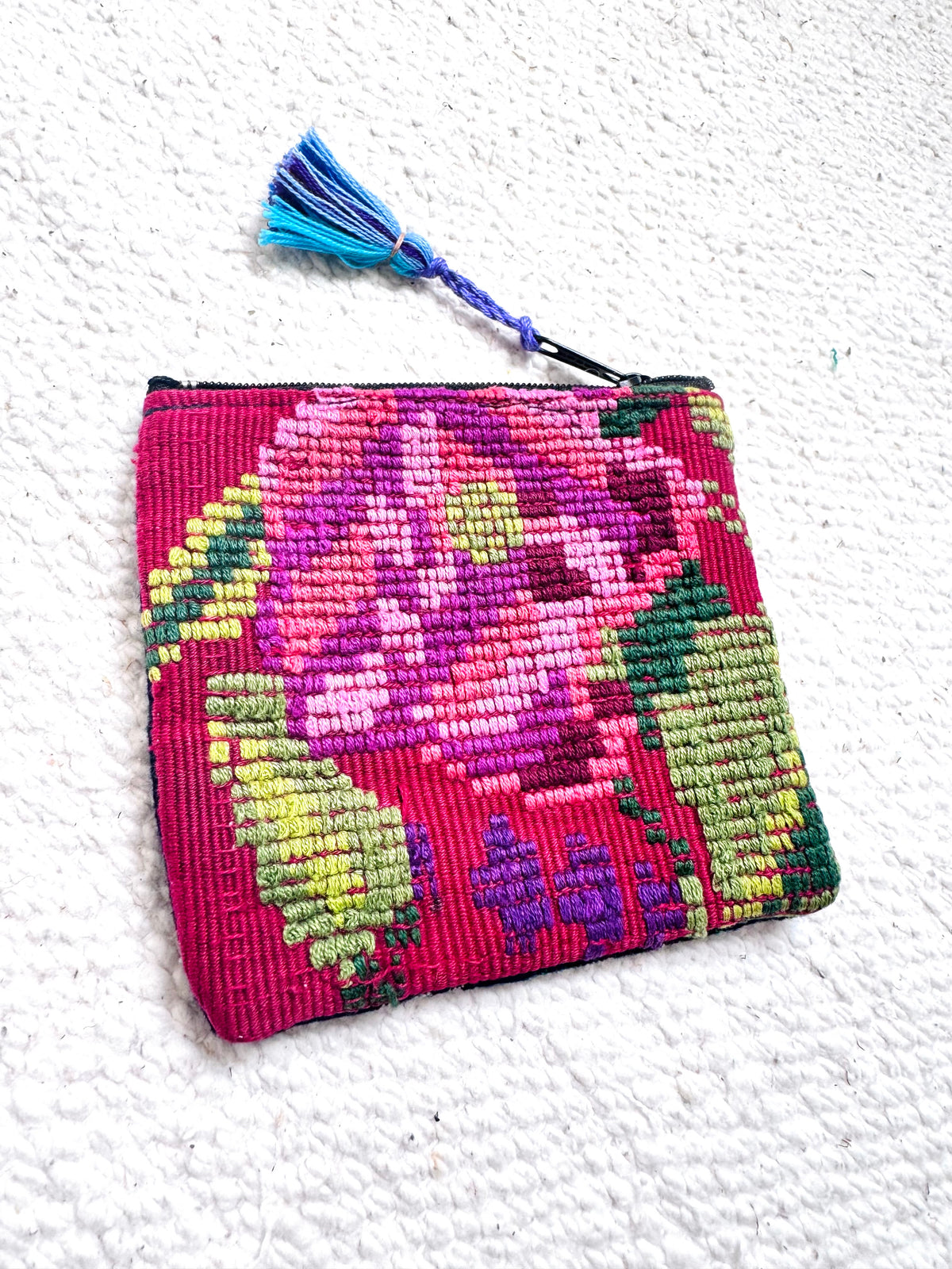 A small rectangular embroidered coin purse with a floral pattern in pink, purple, green, and yellow, featuring a blue and purple tassel zipper pull, set against a textured white background.