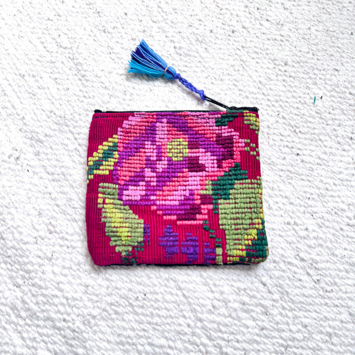 A small rectangular woven pouch with a colorful floral pattern in pink, purple, green, and red, featuring a blue tassel zipper pull, laid flat on a textured white surface.