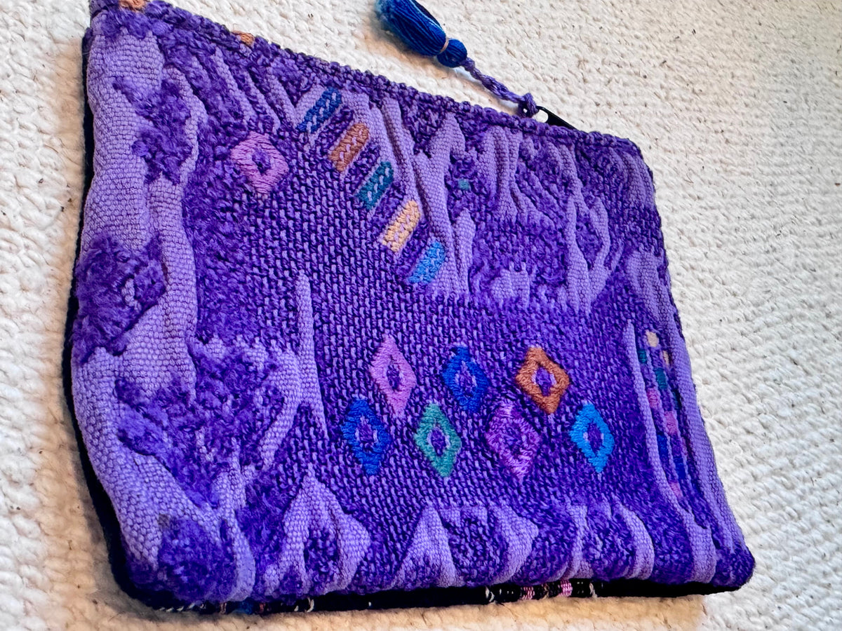 A purple woven clutch bag with a geometric pattern and a blue tassel zipper pull, shown on a textured cream fabric background.
