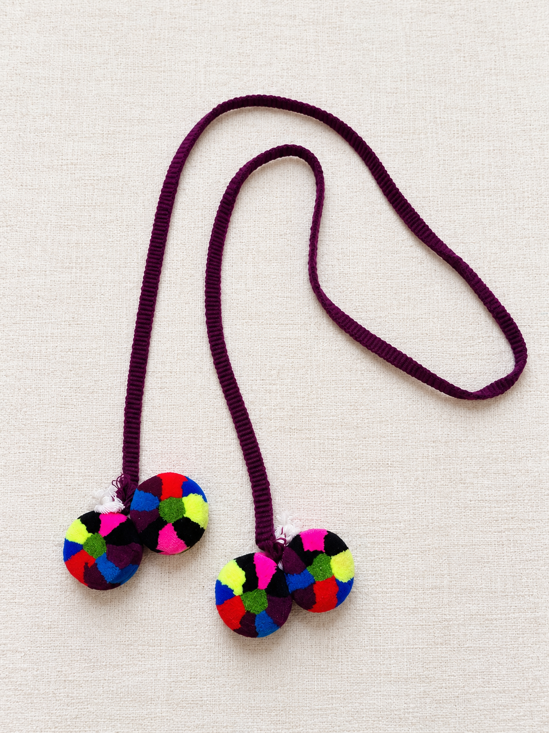 Traditional Chajul hair wrap with a plum-purple cord and four multicolored pom-poms arranged on a cream background