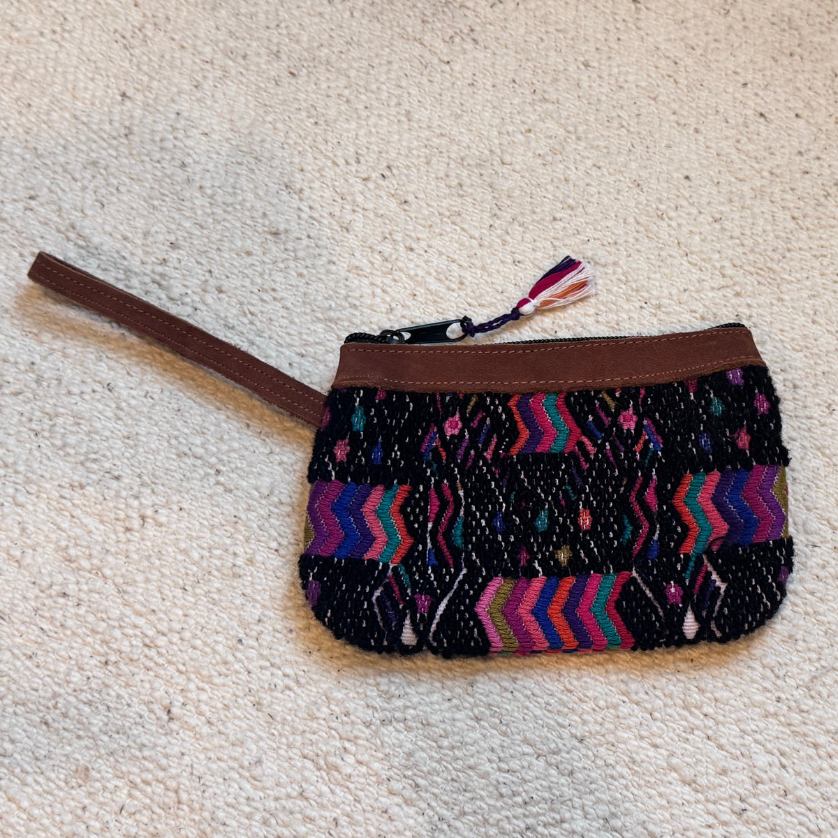 A small woven zip pouch with a multicolor chevron pattern on a black background and brown leather trim, placed on a textured beige carpet.