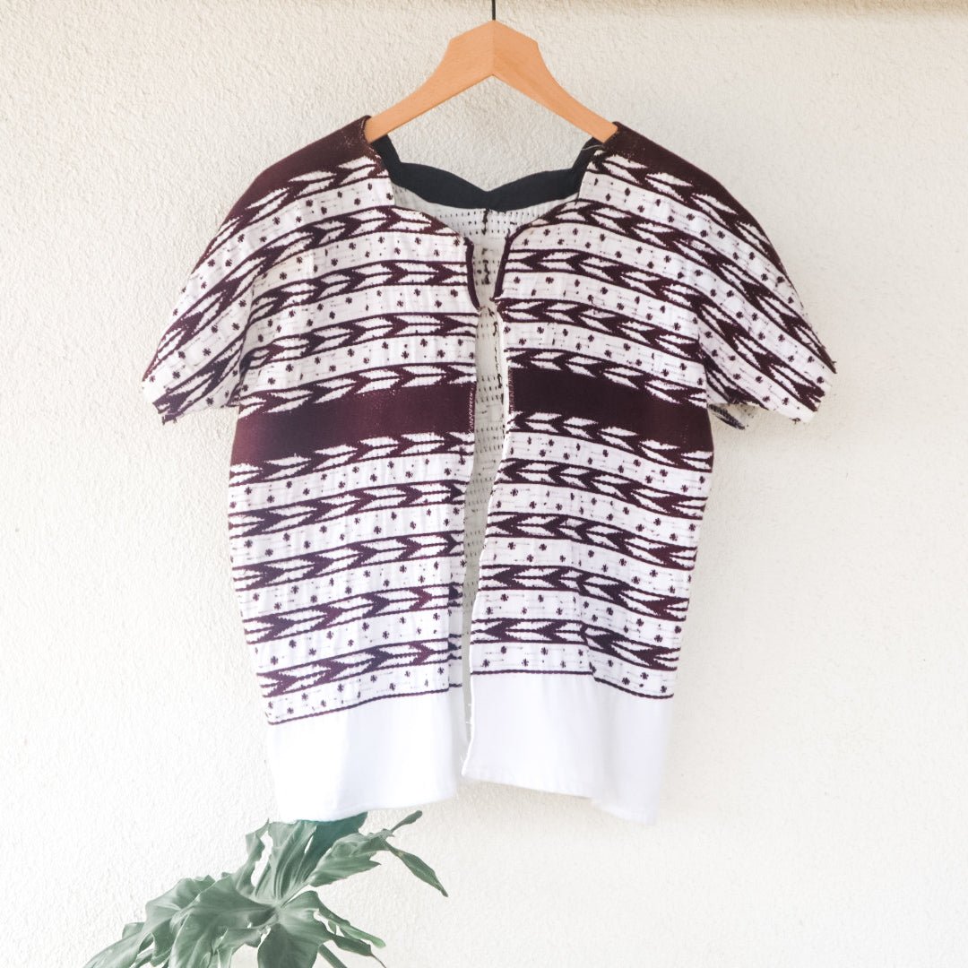 A vintage white Guatemalan Huipil top with geometric brocade patterns, displayed on a hanger against a neutral background with a plant to the side.