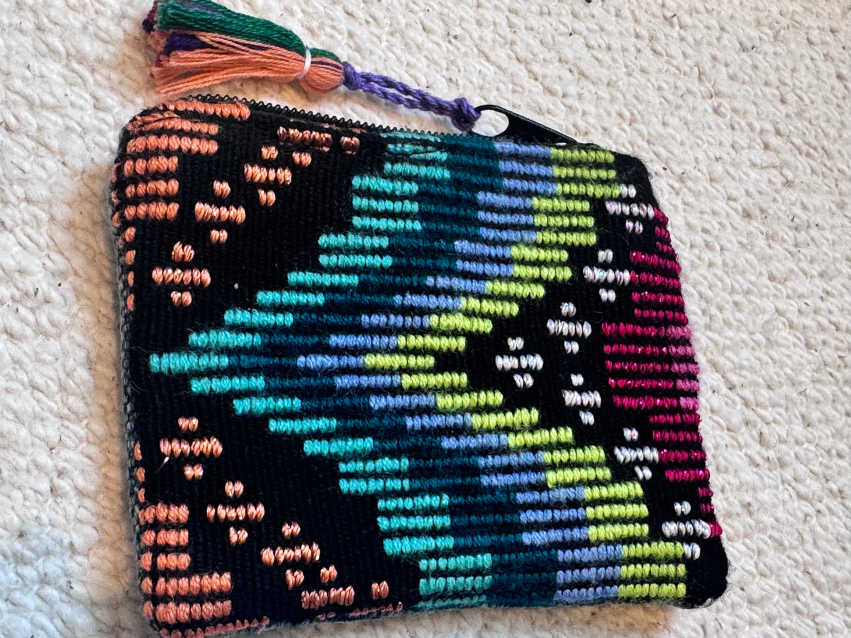 A small zippered pouch with a colorful geometric embroidered pattern on a black background, featuring a tassel on the right side, placed on a textured off-white carpet.