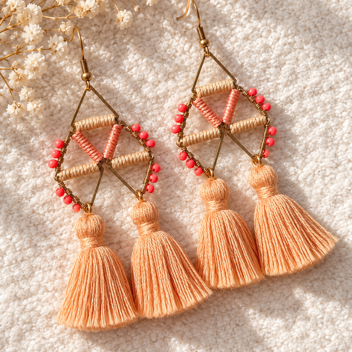 A pair of bohemian-style geometric dangle earrings with peach and coral beads, gold-tone metal frames, and three peach tassels hanging from each earring, displayed on a textured cream fabric background with dried white flowers.