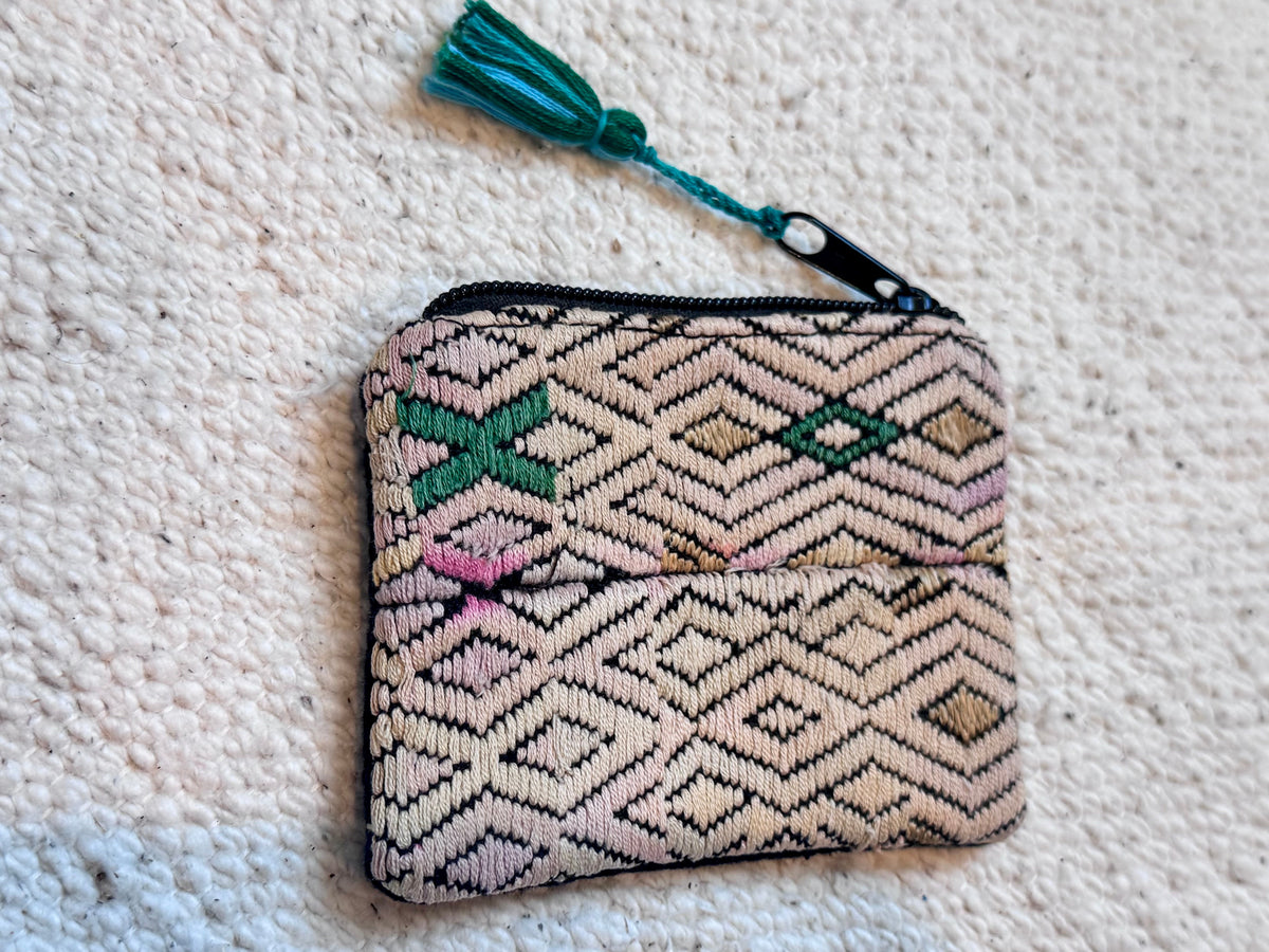 A small woven zip-around pouch with a geometric diamond pattern in beige, brown, and black, accented with pink, green, and beigh, featuring a colorful tassel, laid flat on a textured off-white carpet.