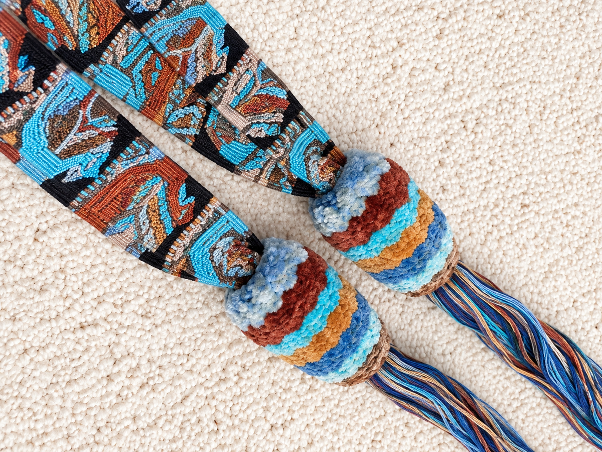 A pair of colorful woven rope accessories with striped knotted loops and patterned hanging strands, laid flat on a textured cream carpet.