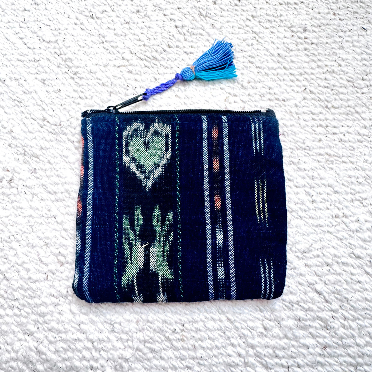 A small woven zip pouch with a dark navy blue base and light blue, white, and mint green geometric patterns, featuring a blue tassel zipper pull, laid flat on a white textured background.
