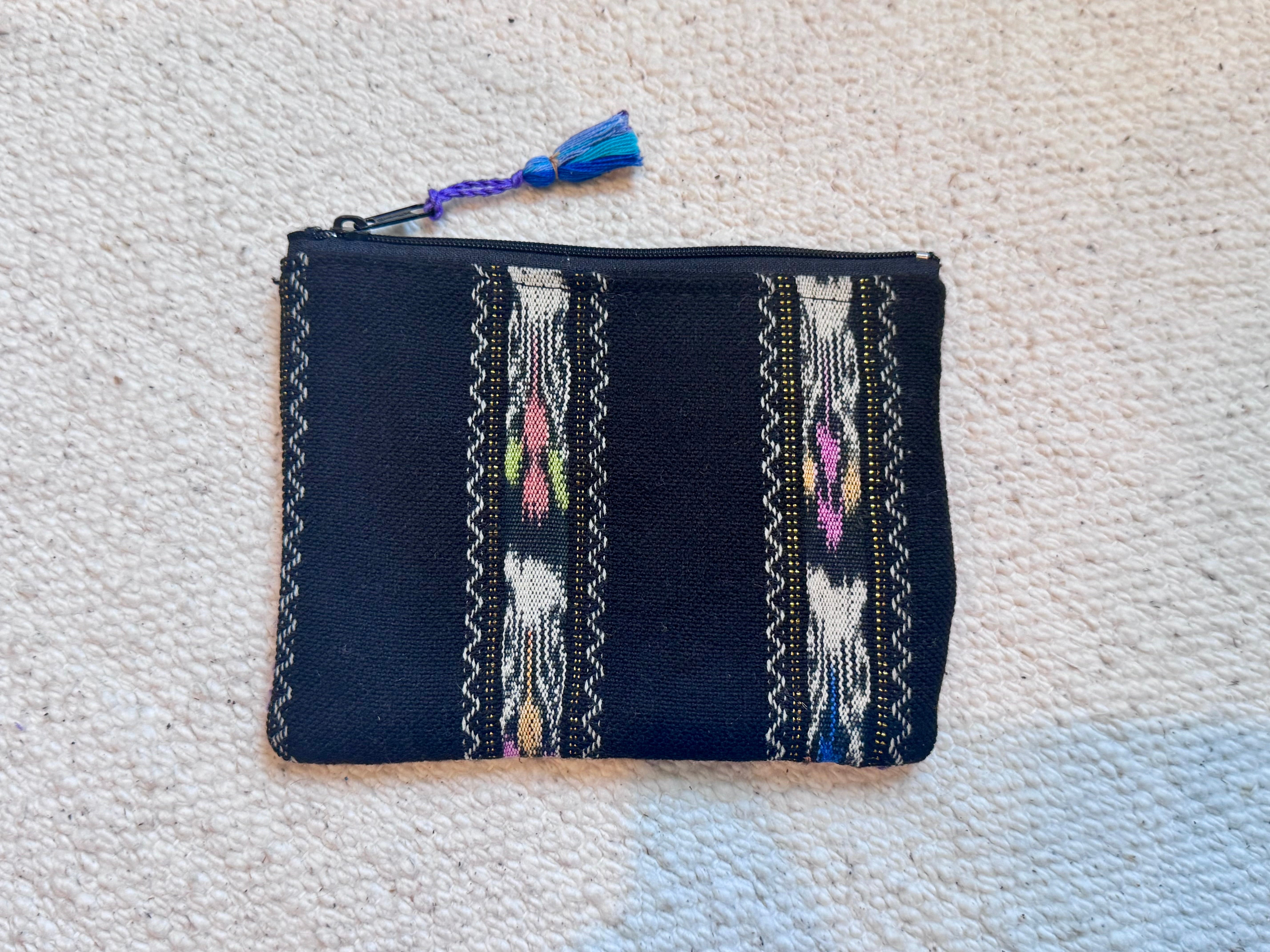 A small zippered pouch or clutch in dark navy fabric with woven geometric and floral patterns in white, pink, yellow, and blue, featuring a blue tassel zipper pull, laid flat on a beige carpet.