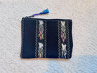 A small zippered pouch or clutch in dark navy fabric with woven geometric and floral patterns in white, pink, yellow, and blue, featuring a blue tassel zipper pull, laid flat on a beige carpet.