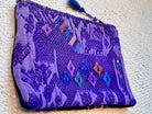 A purple woven clutch bag with a geometric pattern and a blue tassel zipper pull, shown on a textured cream fabric background.