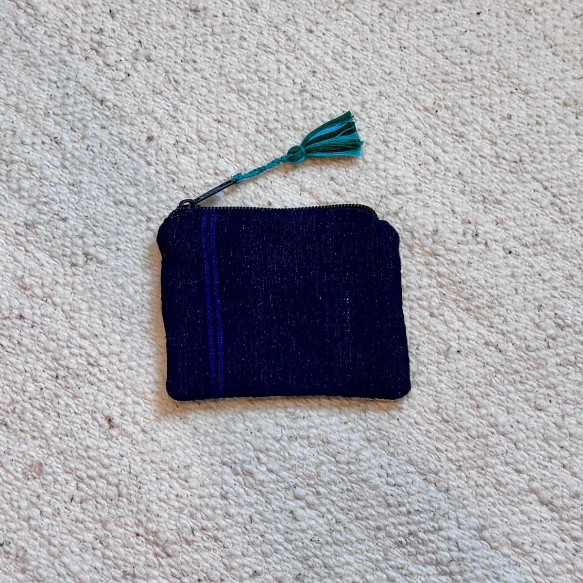 A small dark navy fabric pouch with a blue stripe detail and a teal tassel, placed on a textured off-white carpet.