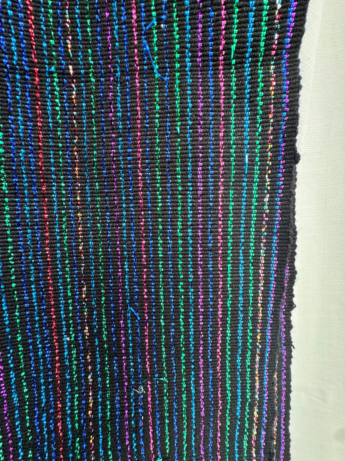 A close-up of a black woven textile with vertical multicolored stripes in blue, green, pink, and purple, next to a plain white background.