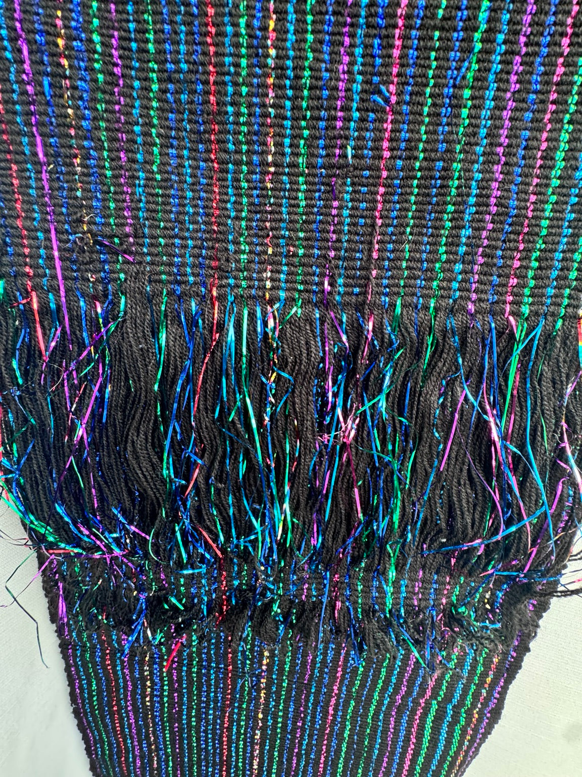 A close-up of a black woven textile with vertical multicolor stripes in blue, green, pink, and purple, featuring a fringed edge.