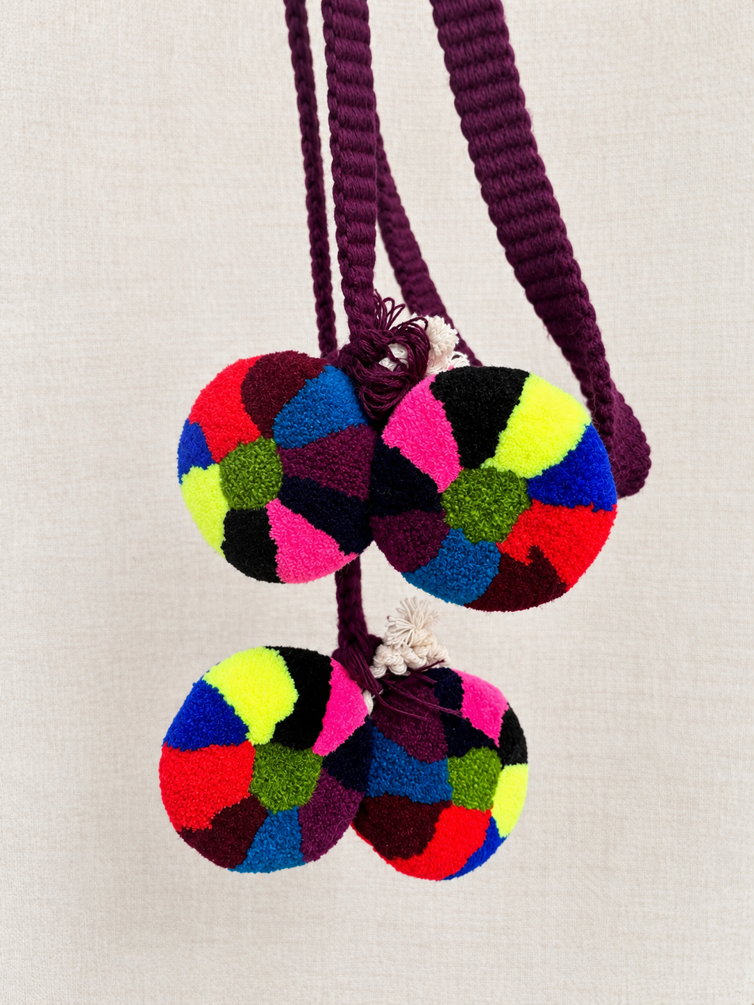 Traditional Chajul hair wrap with a plum-purple cord and four multicolored pom-poms arranged on a cream background. Vida Maya