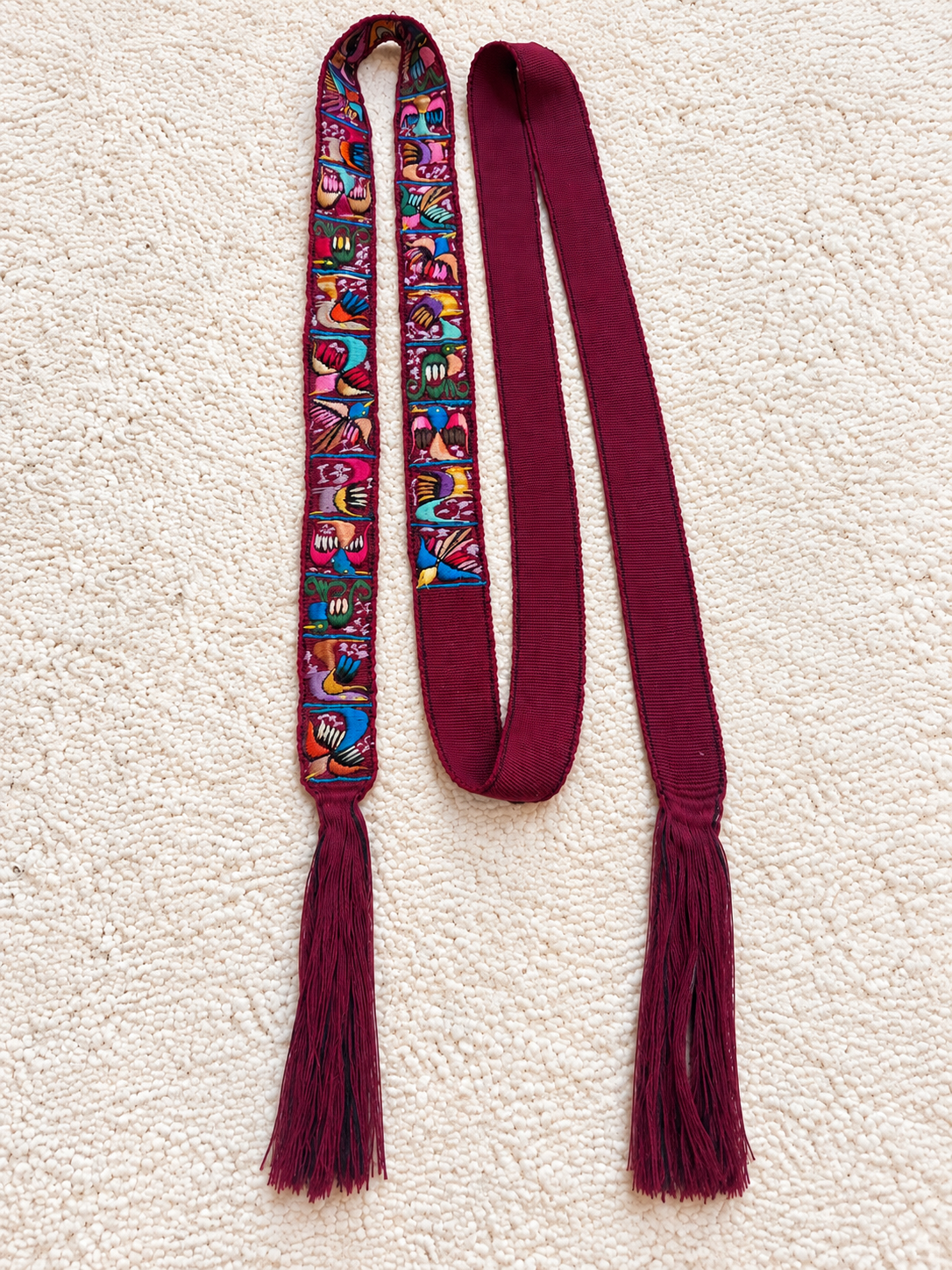 A long scarf laid flat on a textured cream background, featuring a multicolor patterned section with tassels and solid deep burgundy ends and tassels.