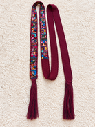 A long scarf laid flat on a textured cream background, featuring a multicolor patterned section with tassels and solid deep burgundy ends and tassels.
