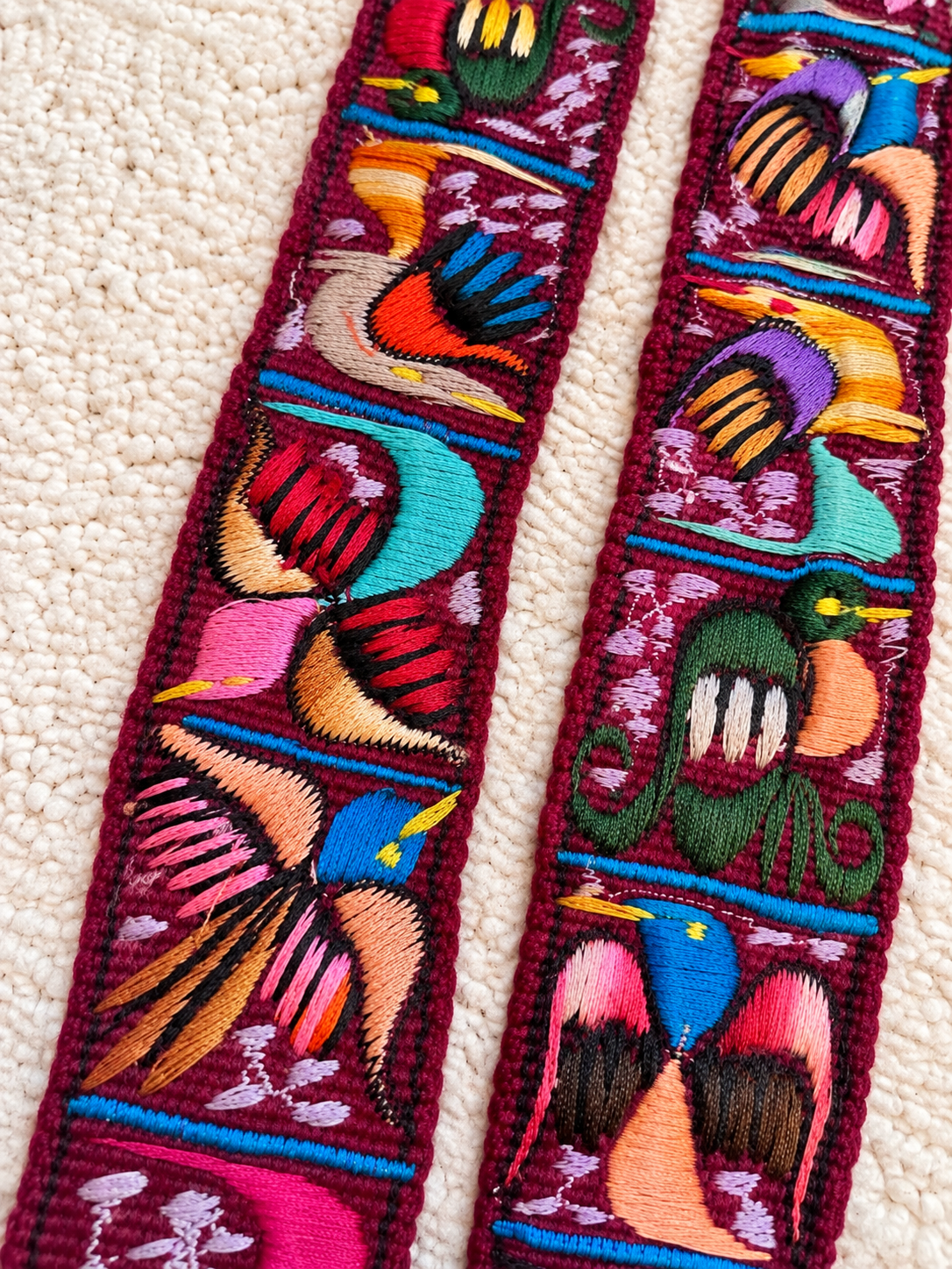 Close-up of two long embroidered decorative straps laid on a cream textured fabric background. The straps feature colorful bird motifs in pink, blue, teal, orange, yellow, green, and purple on a deep burgundy base.