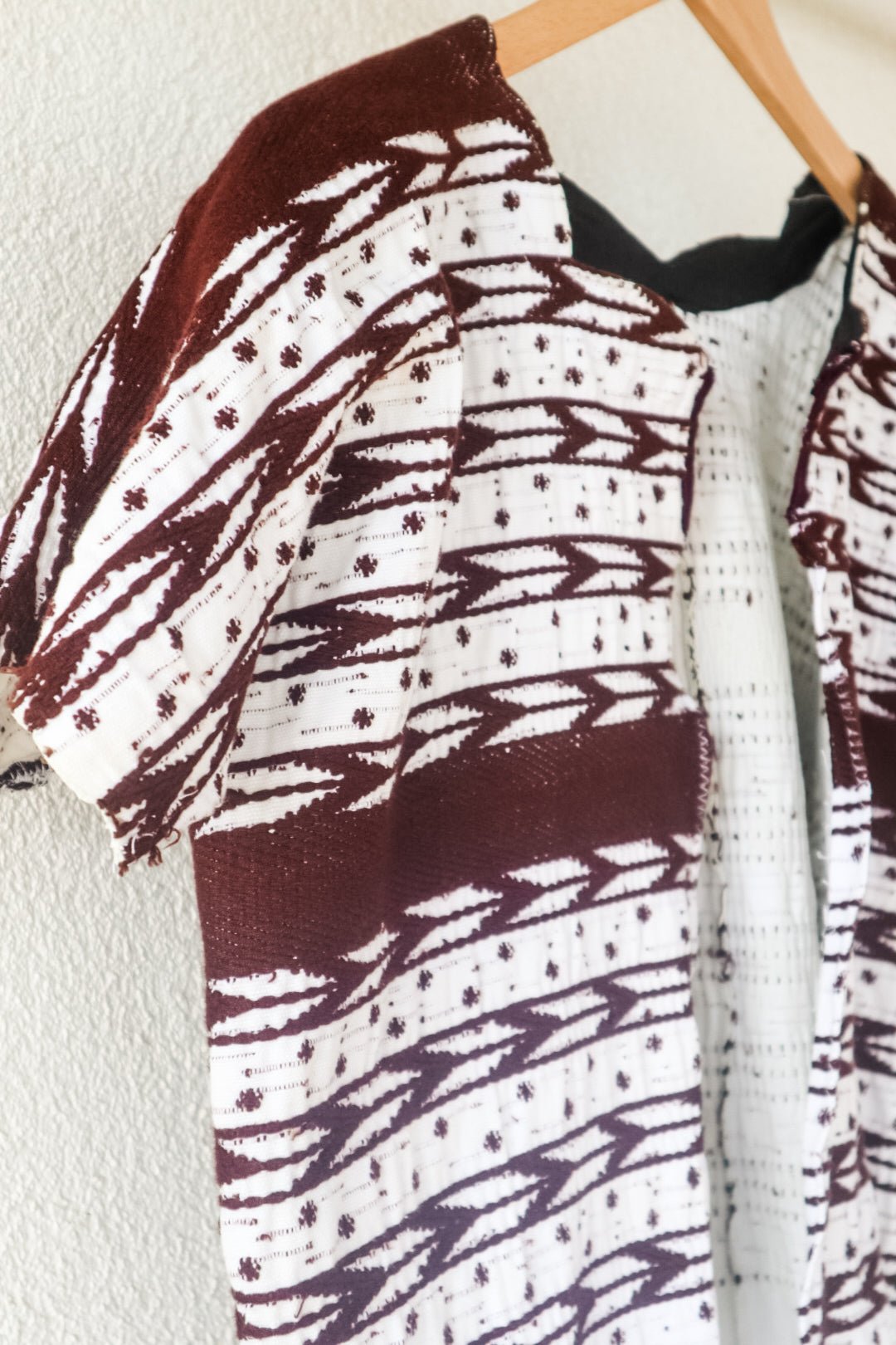 A vintage white Guatemalan Huipil top with geometric brocade patterns, displayed on a hanger against a neutral background with a plant to the side.