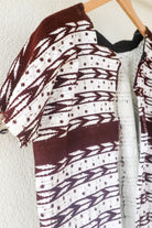 A vintage white Guatemalan Huipil top with geometric brocade patterns, displayed on a hanger against a neutral background with a plant to the side.