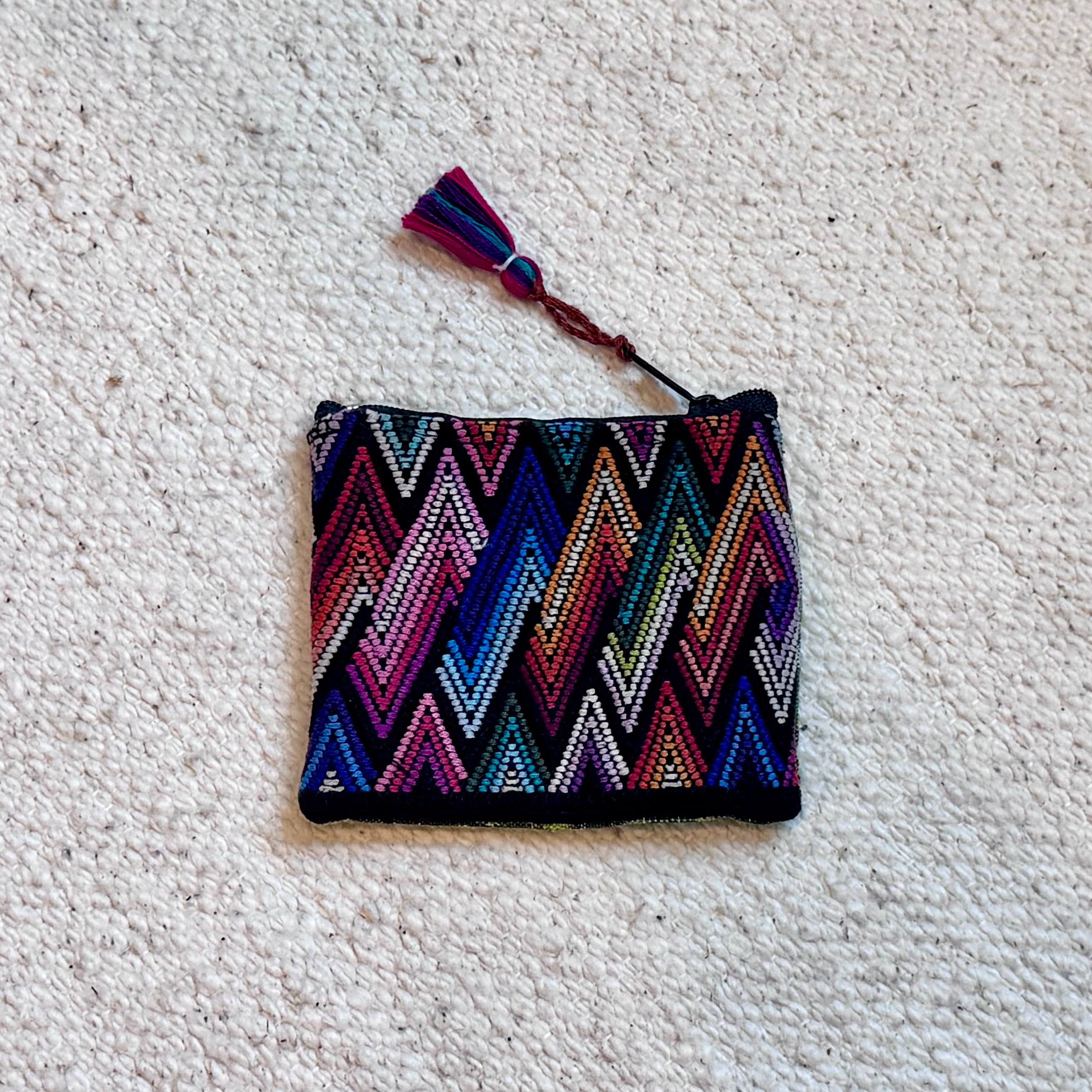 A small woven zip pouch with a colorful chevron pattern in pink, blue, purple, and white, featuring a matching tassel, placed on a textured off-white carpet.