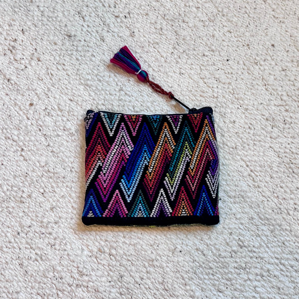 A small woven zip pouch with a colorful chevron pattern in pink, blue, purple, and white, featuring a matching tassel, placed on a textured off-white carpet.