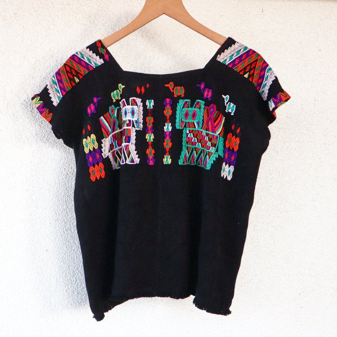 A vintage black Huipil top with colorful geometric embroidery, hanging on a wooden hanger.