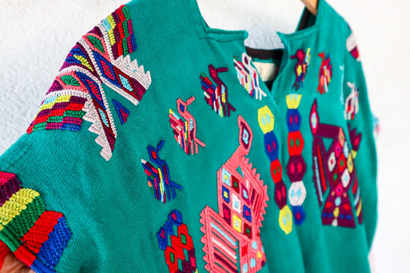 A vintage Mayan Huipil top with embroidered peacock and butterfly designs on a dark teal background, displayed on a wooden hanger.