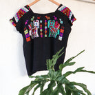 Black Guatemalan huipil hanging against a white wall with colorful woven motifs and green leaves in the foreground