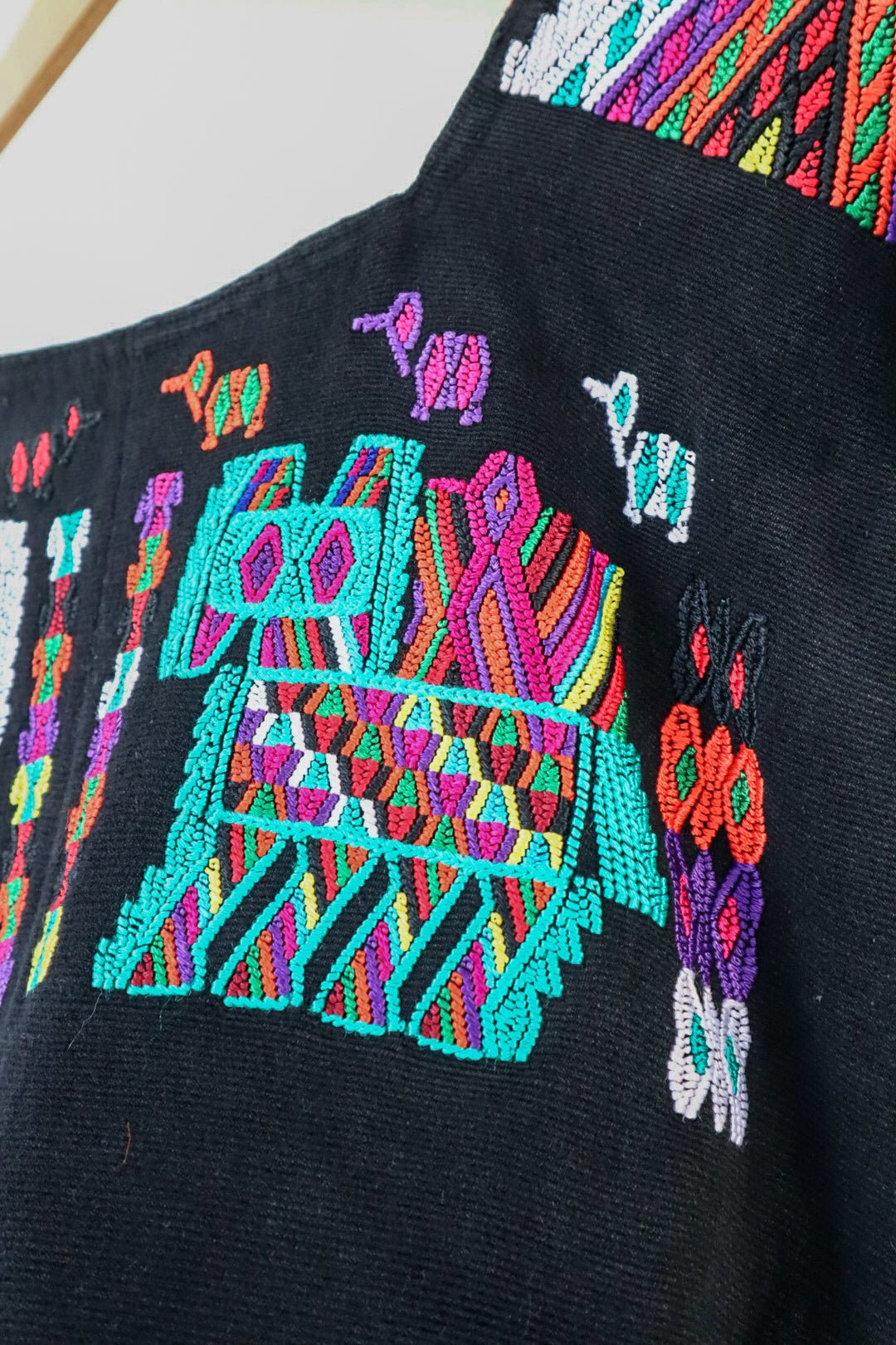 Black Guatemalan huipil hanging against a white wall with colorful woven motifs and green leaves in the foreground