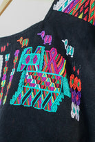 Black Guatemalan huipil hanging against a white wall with colorful woven motifs and green leaves in the foreground