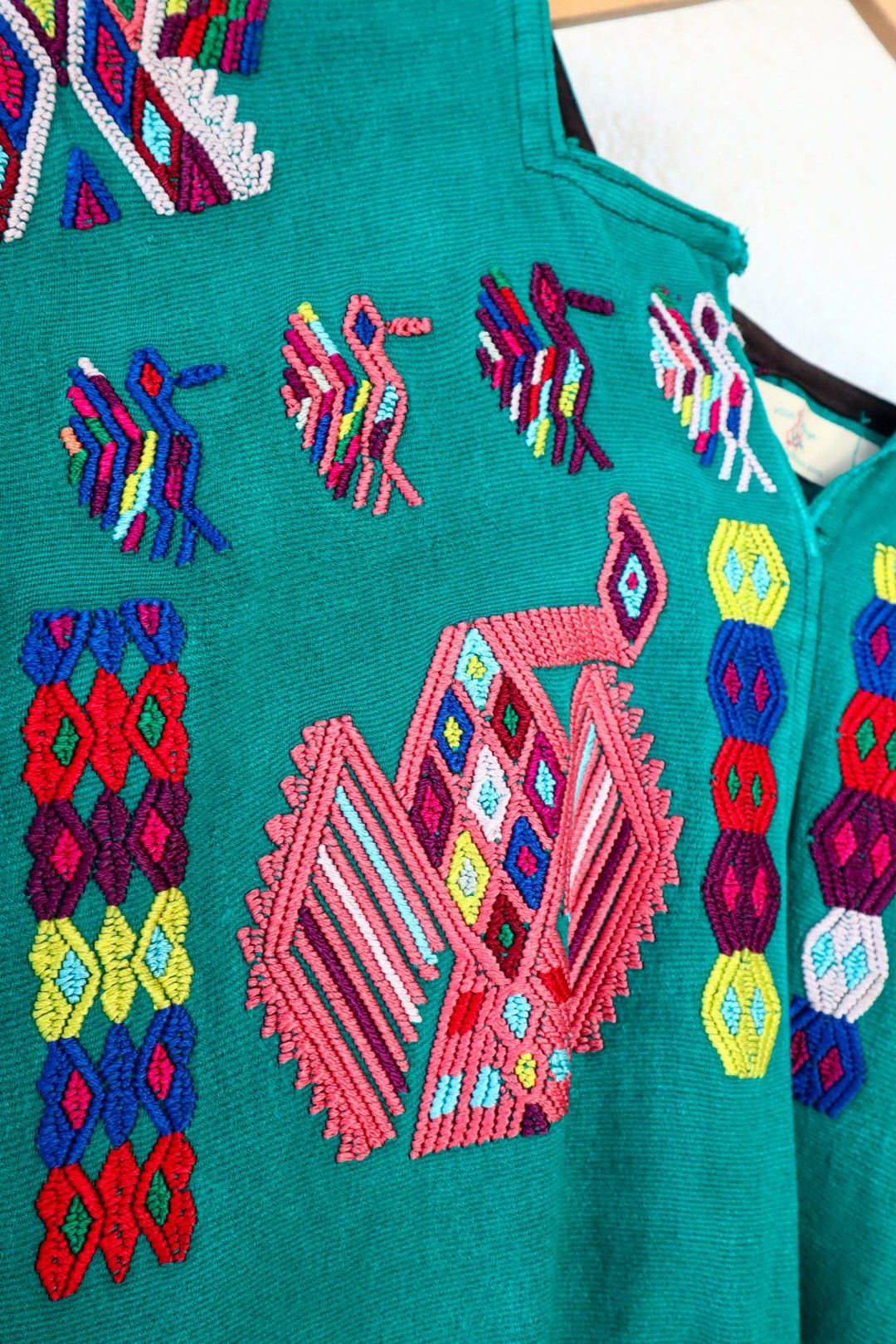 A vintage Mayan Huipil top with embroidered peacock and butterfly designs on a dark teal background, displayed on a wooden hanger.