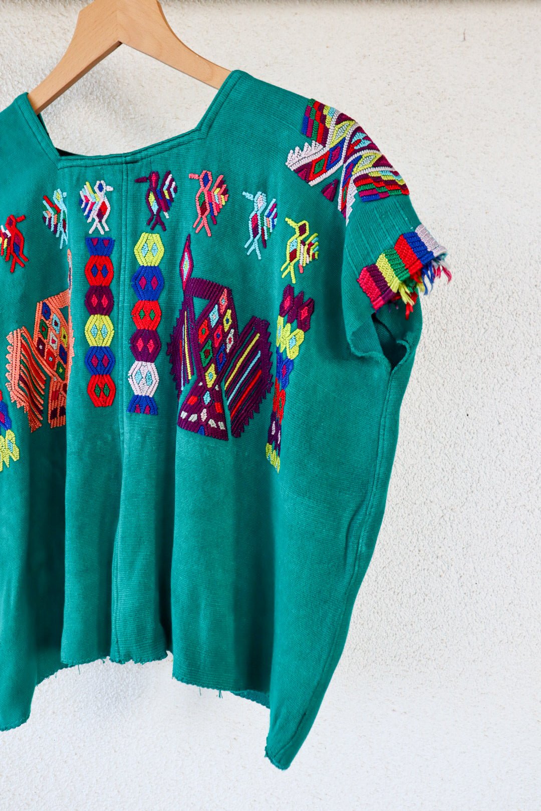 A vintage Mayan Huipil top with embroidered peacock and butterfly designs on a dark teal background, displayed on a wooden hanger.