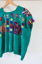 A vintage Mayan Huipil top with embroidered peacock and butterfly designs on a dark teal background, displayed on a wooden hanger.