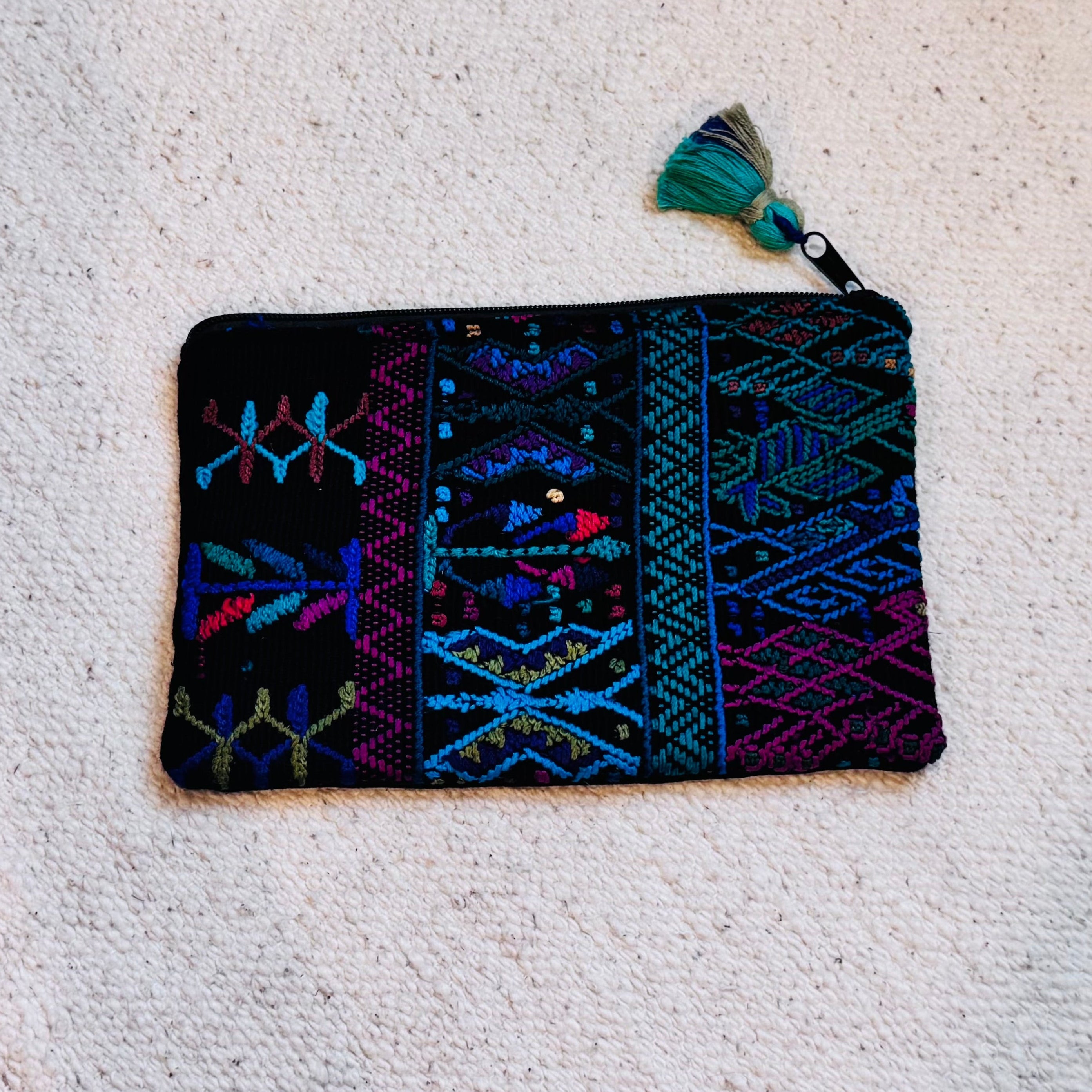 A rectangular black woven pouch with colorful geometric embroidery in blue, purple, and green, featuring a matching tassel zipper pull, laid flat on a textured off-white carpet.
