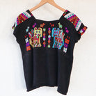 Black Guatemalan huipil hanging against a white wall with colorful woven motifs and green leaves in the foreground
