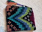 A small zippered pouch with a colorful geometric embroidered pattern on a black background, featuring a tassel on the right side, placed on a textured off-white carpet.