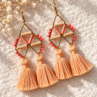 A pair of bohemian-style geometric dangle earrings with peach and coral beads, gold-tone metal frames, and three peach tassels hanging from each earring, displayed on a textured cream fabric background with dried white flowers.
