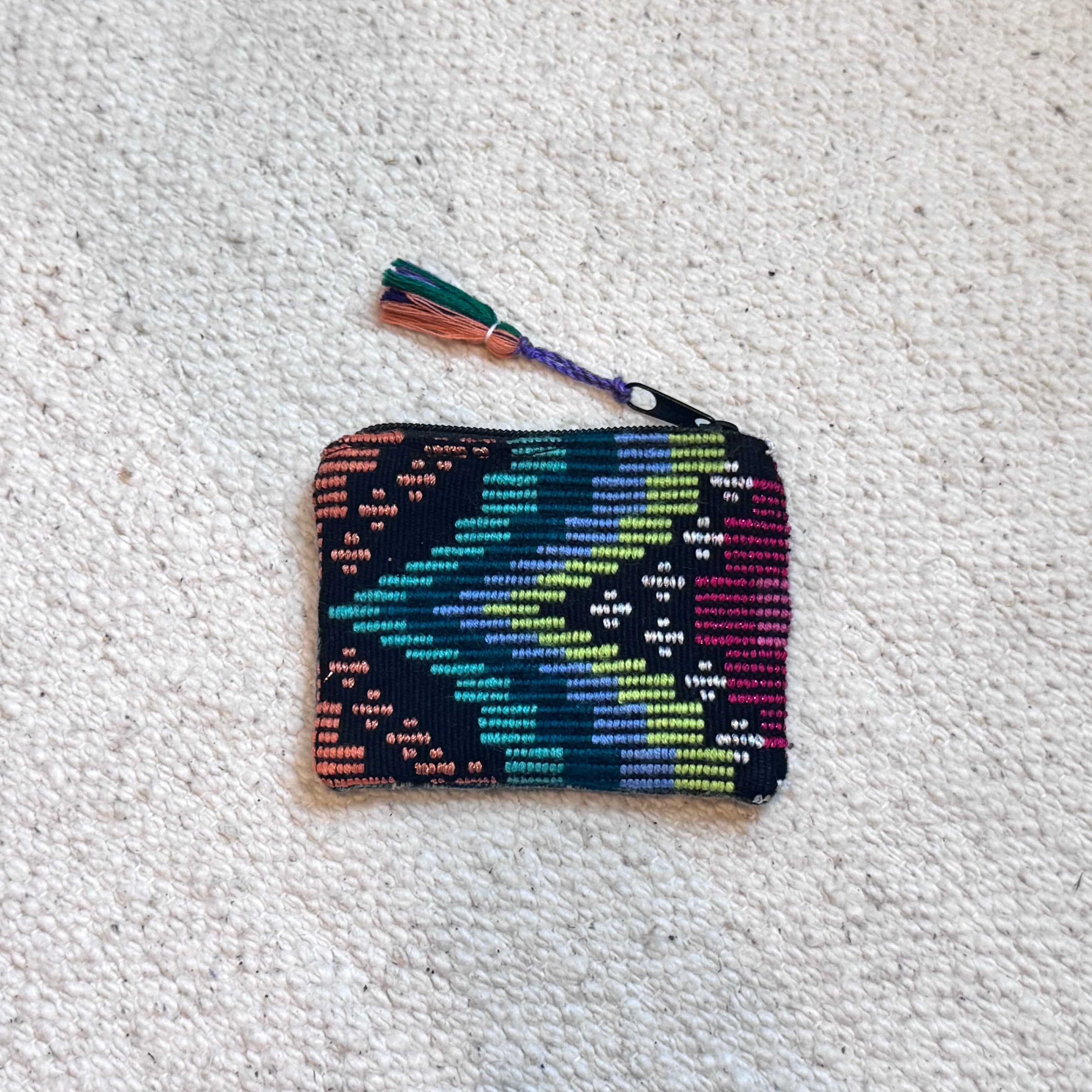 A small woven zip pouch with a colorful geometric pattern in teal, green, yellow, orange, and purple, featuring a matching tassel, laid flat on a textured off-white carpet.