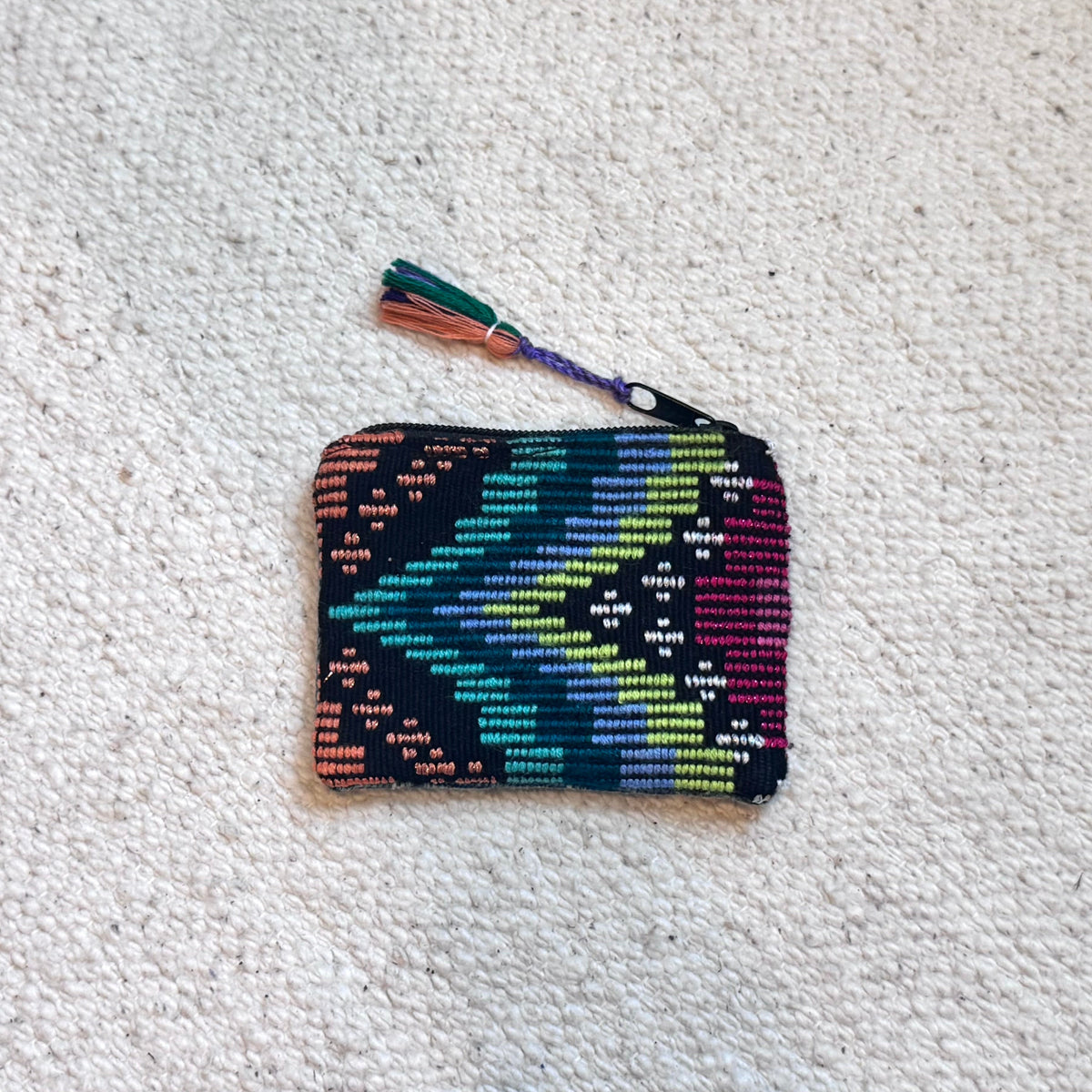 A small woven zip pouch with a colorful geometric pattern in teal, green, yellow, orange, and purple, featuring a matching tassel, laid flat on a textured off-white carpet.