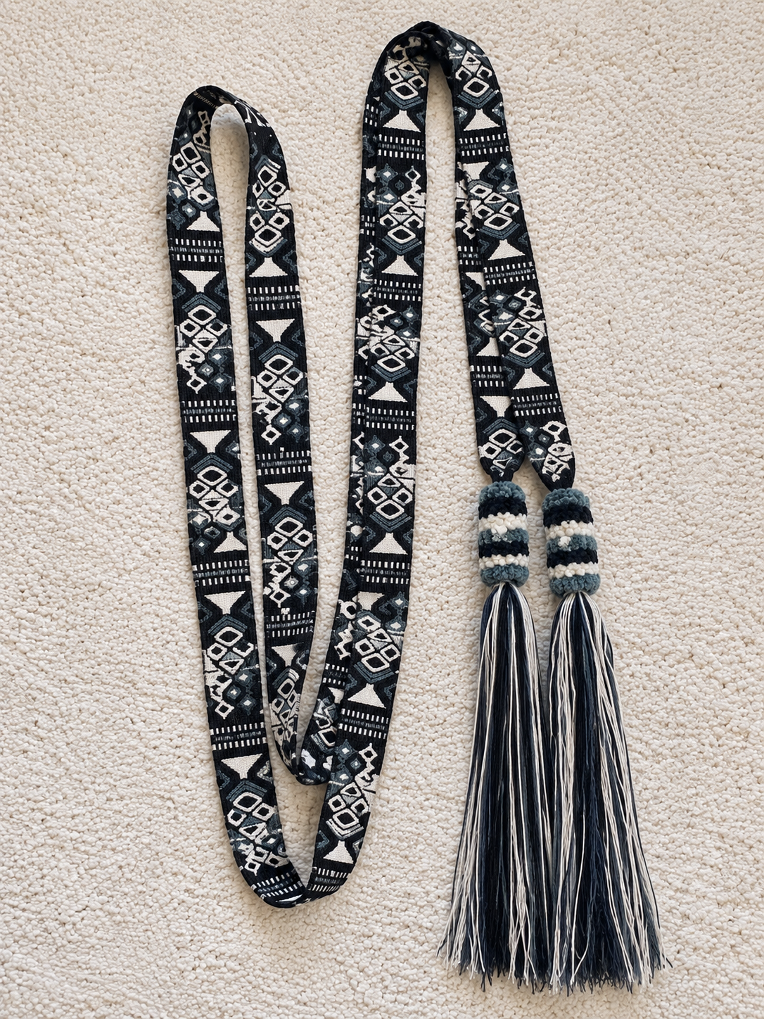 A long woven scarf laid flat on a textured cream fabric background. The scarf has a black and white geometric pattern with two knotted tassel ends in black, white, and teal-blue.