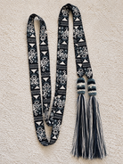 A long woven scarf laid flat on a textured cream fabric background. The scarf has a black and white geometric pattern with two knotted tassel ends in black, white, and teal-blue.