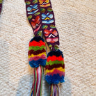 A close-up of a colorful woven textile accessory laid on a beige carpet, featuring a dark patterned strap with bright multicolored motifs and two fuzzy tassels in rainbow stripes.