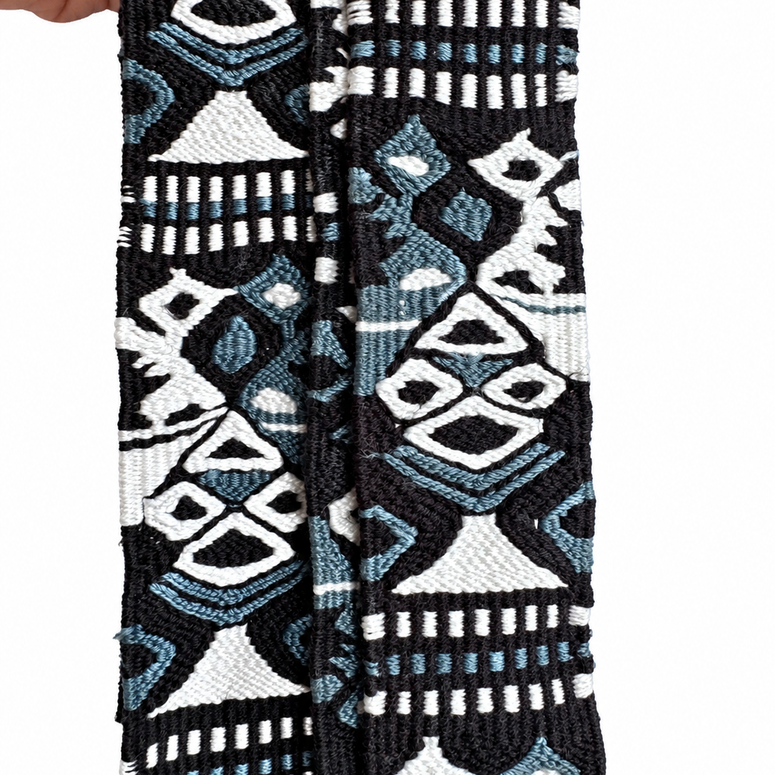 A close-up of a woven textile strap or belt with a geometric tribal pattern in black, white, and light blue, held by a hand on the left side against a white background.