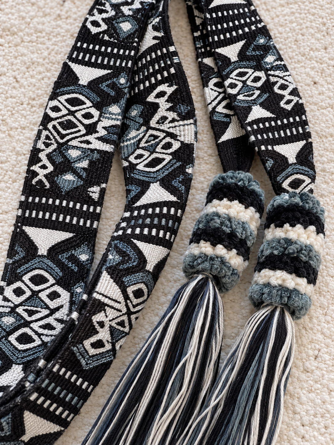 A close-up of a woven textile accessory, likely a belt or scarf, featuring a black, white, and gray geometric pattern with two knotted ends wrapped in striped yarn and finished with long tassels, laid flat on a beige textured surface.