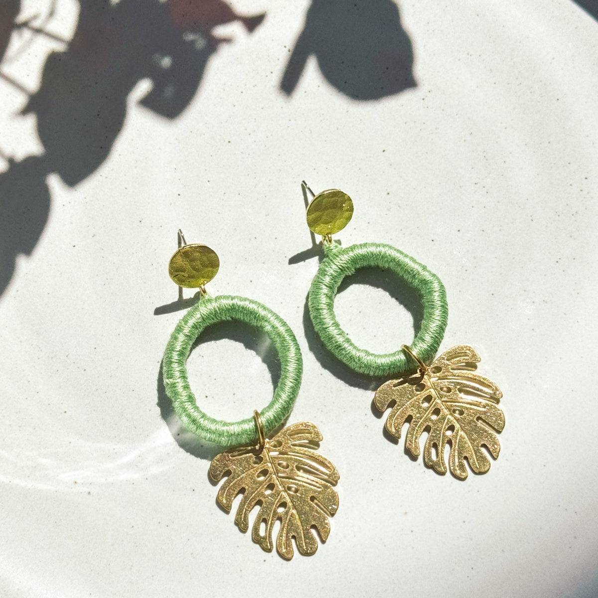 A pair of green earrings with gold-colored hooks, designed to resemble tropical leaves, placed on a white plate with a shadow of a plant in the background.