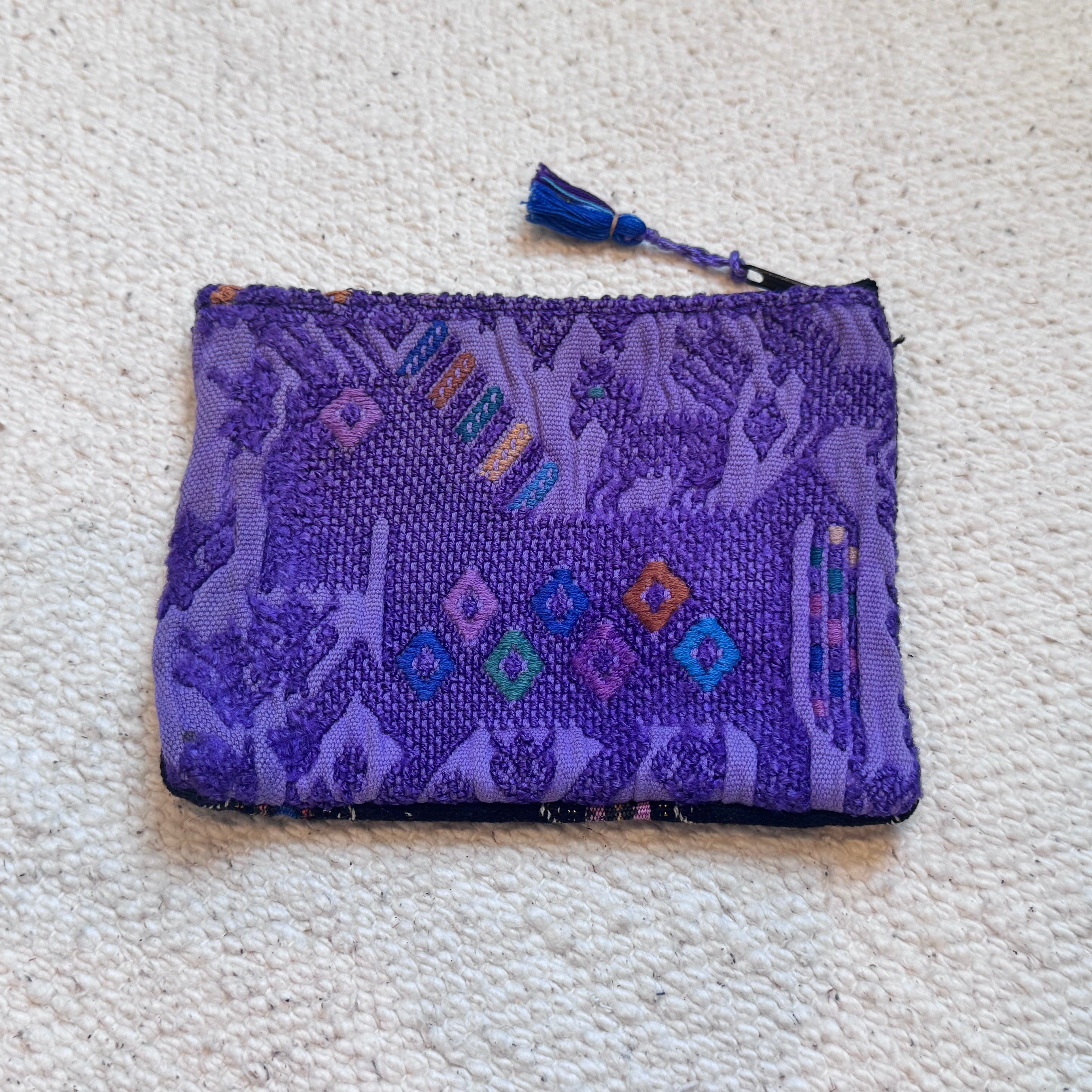 A purple woven zip pouch with a blue tassel zipper pull, featuring a batik-style pattern of deer and colorful embroidered diamond accents, laid flat on a textured off-white carpet.