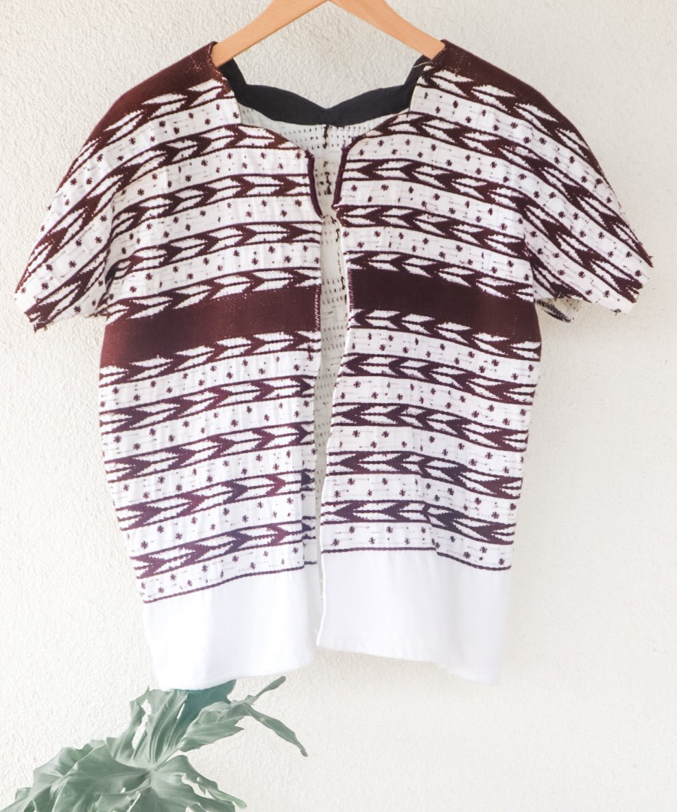 A vintage white Guatemalan Huipil top with geometric brocade patterns, displayed on a hanger against a neutral background with a plant to the side.