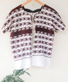 A vintage white Guatemalan Huipil top with geometric brocade patterns, displayed on a hanger against a neutral background with a plant to the side.
