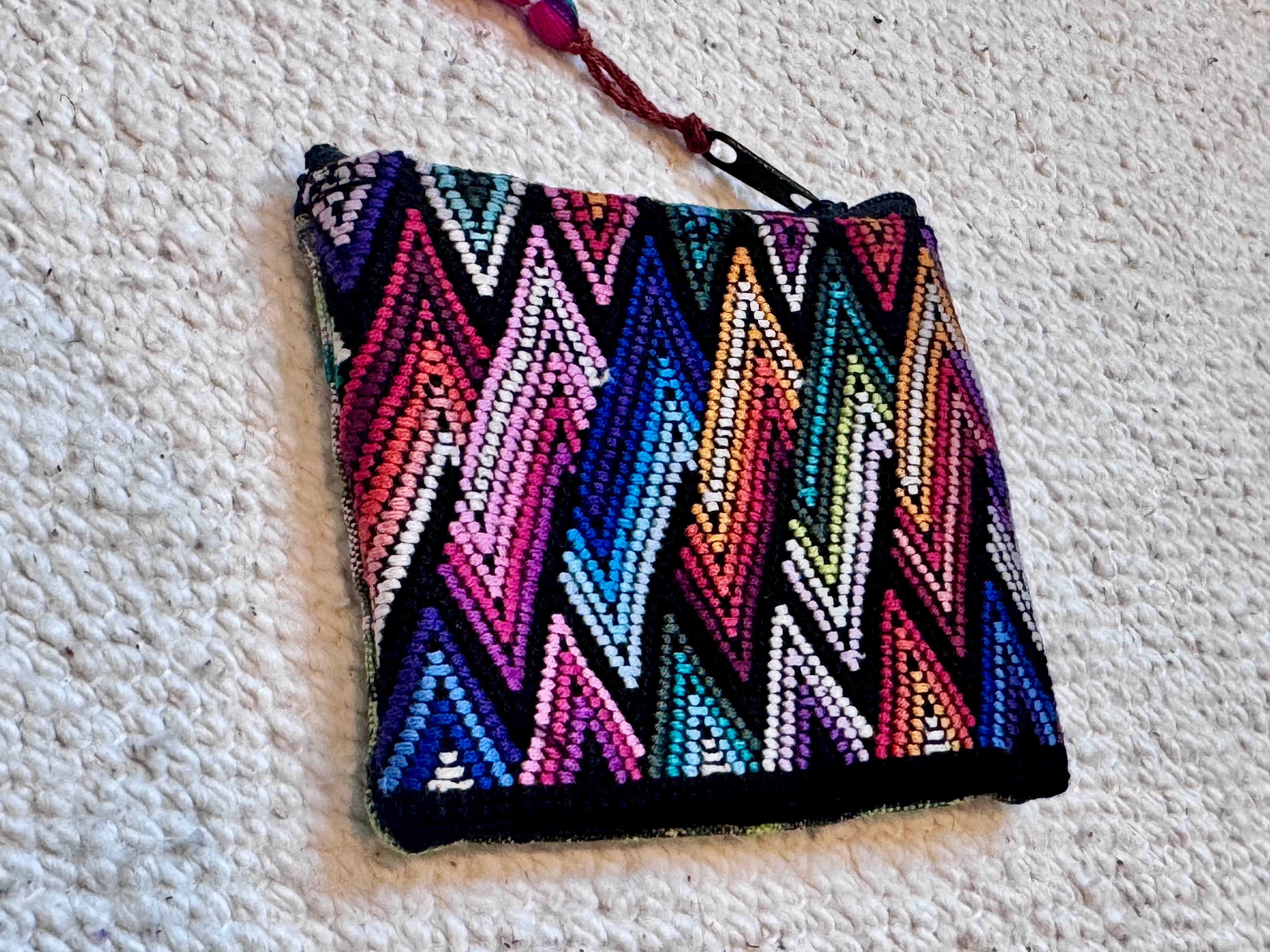 A small zippered pouch with a vibrant multicolor chevron pattern on a black background, featuring a tassel zipper pull, photographed on a textured white knit blanket.