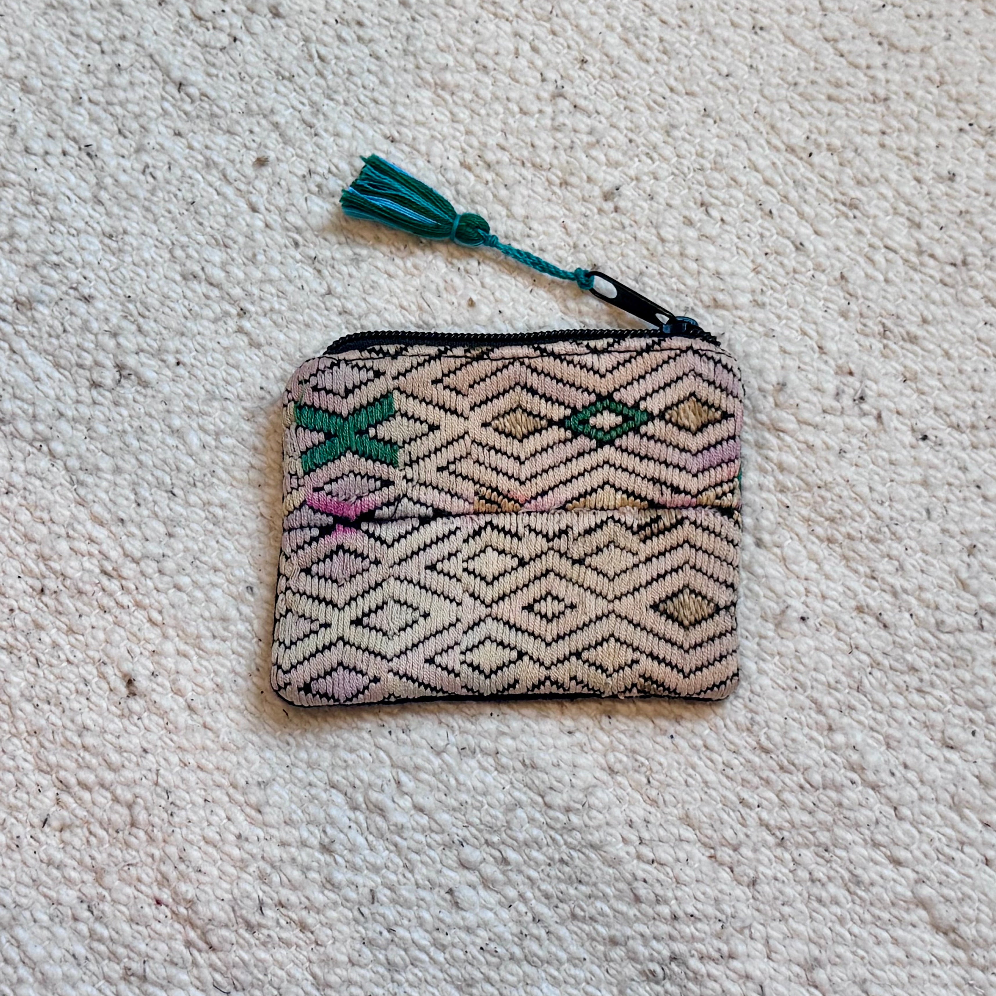 A small woven zip-around pouch with a geometric diamond pattern in beige, brown, and black, accented with pink, green, and beigh, featuring a colorful tassel, laid flat on a textured off-white carpet.